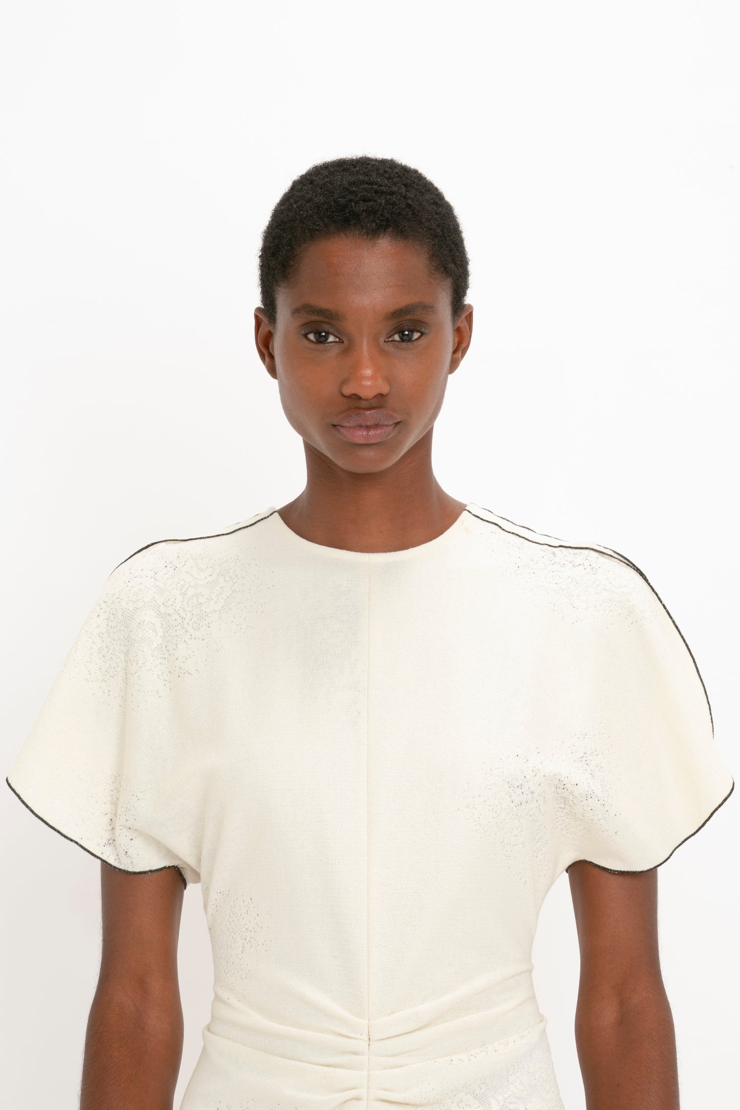 A young black woman wearing a Victoria Beckham Gathered Waist Midi Dress In Cream stands against a white background, looking directly at the camera.