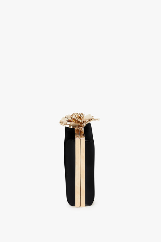 A black and gold cylindrical gift box tied with a golden ribbon and bow, featuring a hand-crafted Victoria Beckham Frame Flower Minaudiere in Black, isolated on a white background.