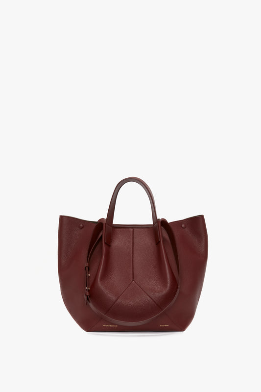 A Victoria Beckham Medium Tote In Burgundy Leather handbag with a pentagonal design at center, dual handles, and visible stitching, displayed against a white background.
