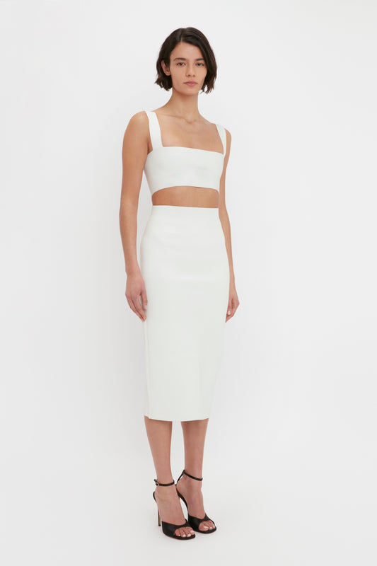 A woman models a white two-piece dress featuring a VB Body Strap Bandeau top and knee-length skirt, paired with black heels, standing against a plain white background from Victoria Beckham.