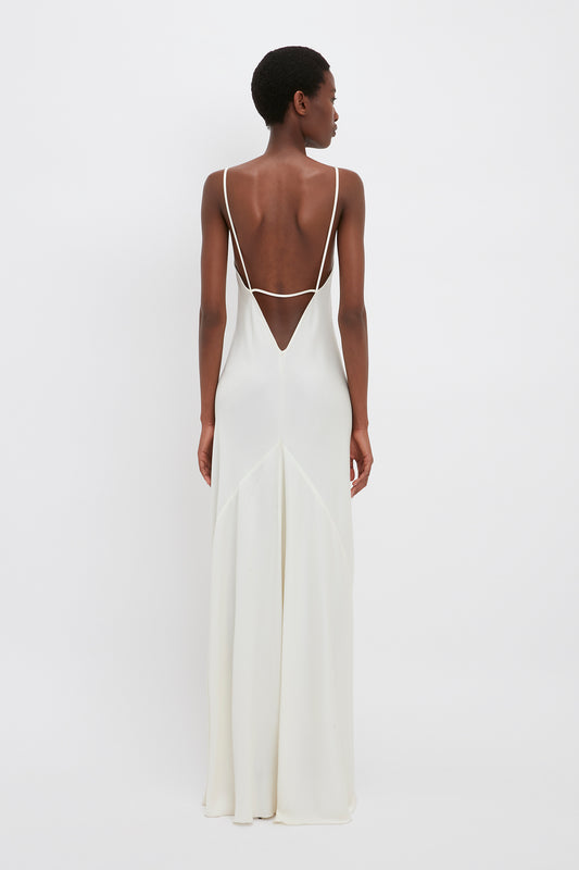 A woman standing with her back to the camera, wearing an elegant Victoria Beckham floor-length cami dress in ivory with a plunging back and thin straps.