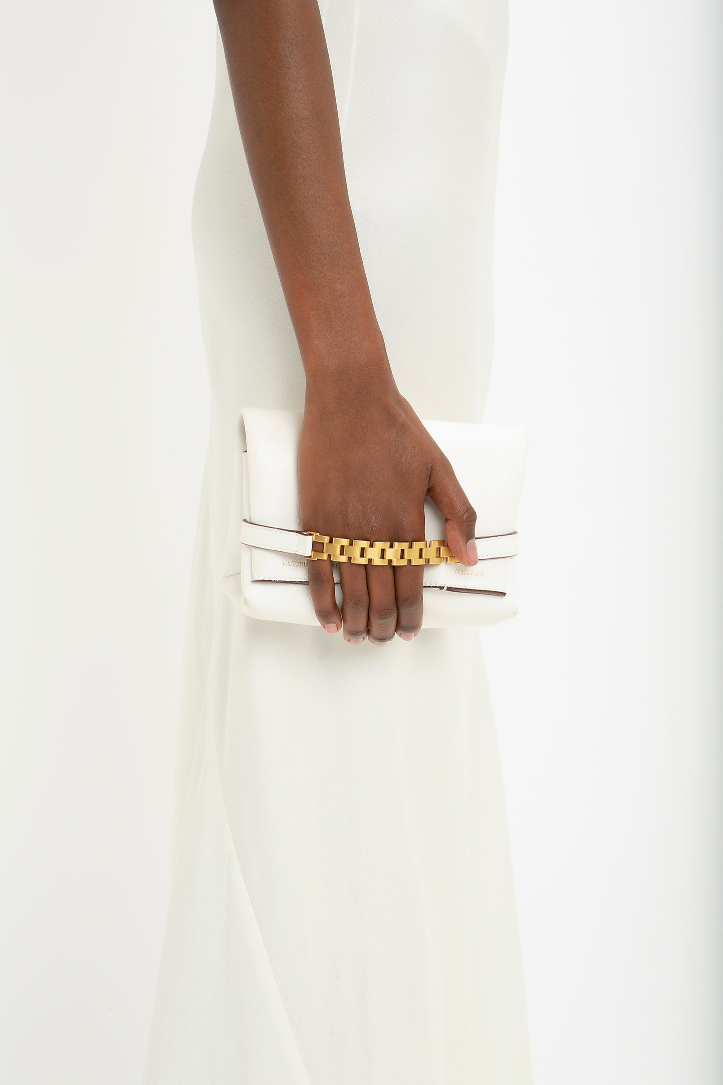A woman's hand holding a white clutch bag with a gold chain, wearing a Victoria Beckham floor-length cami dress in ivory.