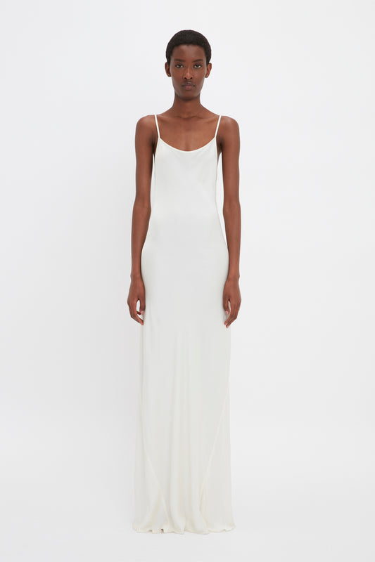 A black woman in a Victoria Beckham floor-length cami dress in ivory stands against a plain white background, looking directly at the camera.