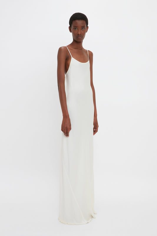 A slender woman in a long, elegant Victoria Beckham floor-length cami dress in ivory standing against a white background.