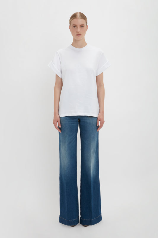 A woman standing in a plain white studio background, wearing Victoria Beckham's Asymmetric Relaxed Fit T-Shirt In White and wide-legged blue jeans.