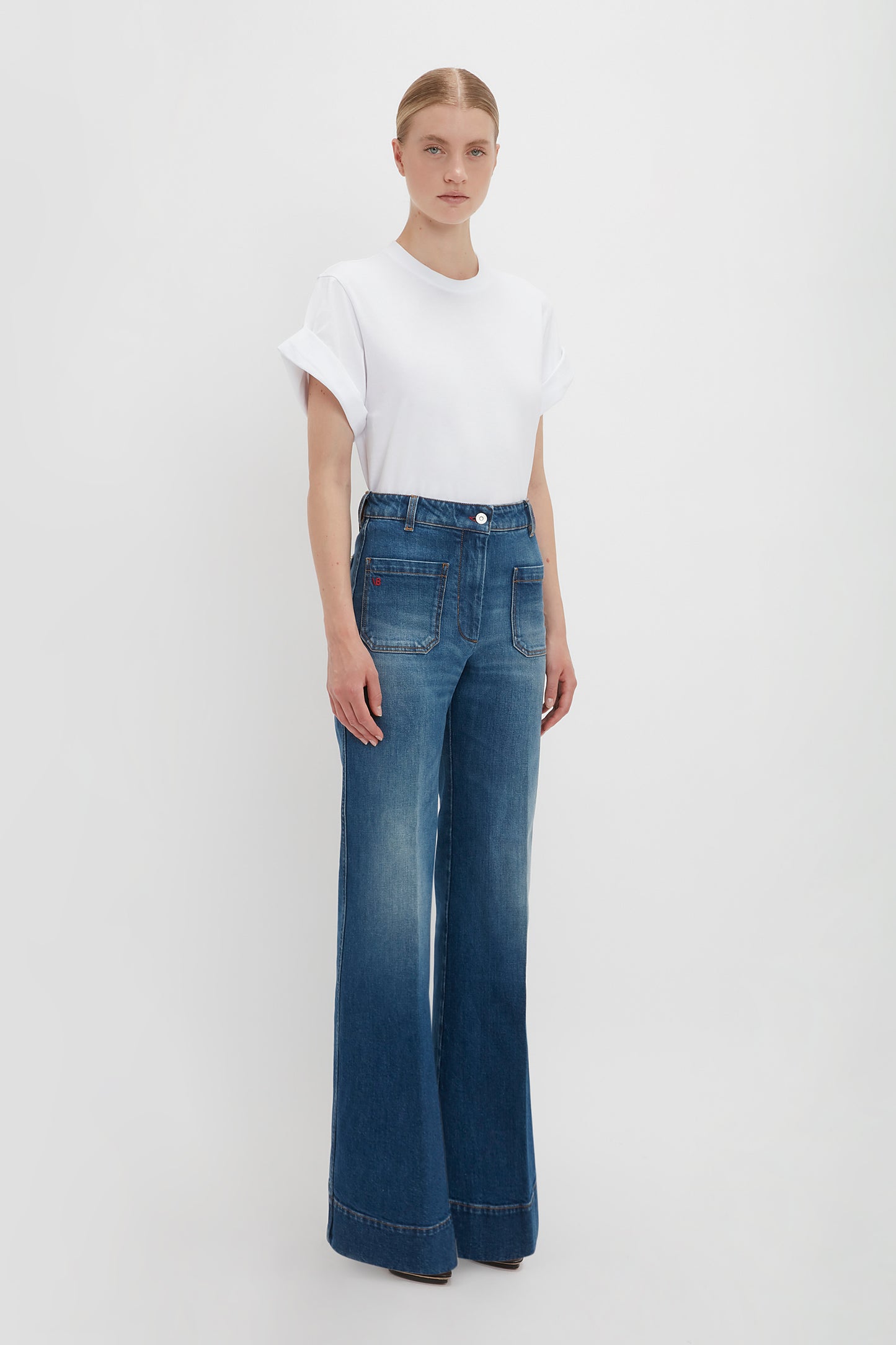 A woman stands facing forward wearing an oversized white Victoria Beckham T-shirt and blue high-waisted flared jeans in dark vintage wash against a plain white background.