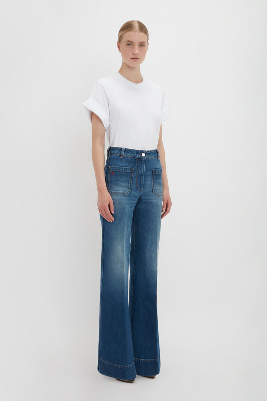 A woman stands facing forward wearing an oversized white Victoria Beckham T-shirt and blue high-waisted flared jeans in dark vintage wash against a plain white background.