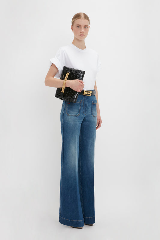 A woman in an oversized Asymmetric Relaxed Fit T-Shirt in White and Alina jeans in dark vintage wash holding a black clutch and wearing a gold belt, standing against a white background by Victoria Beckham.