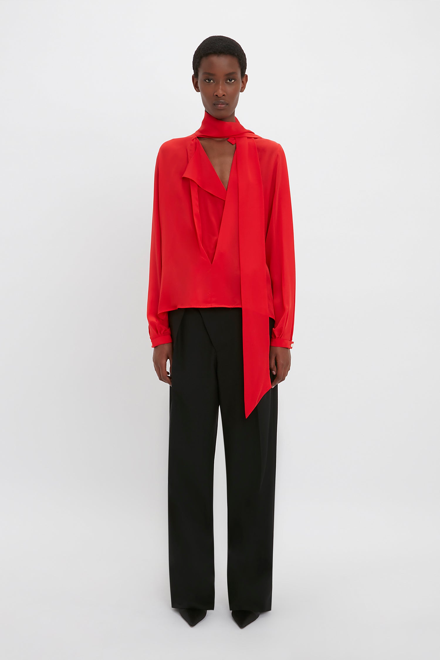 Person stands against a plain background wearing the Victoria Beckham Scarf Neck Blouse In Bright Red, paired with black wide-legged trousers.
