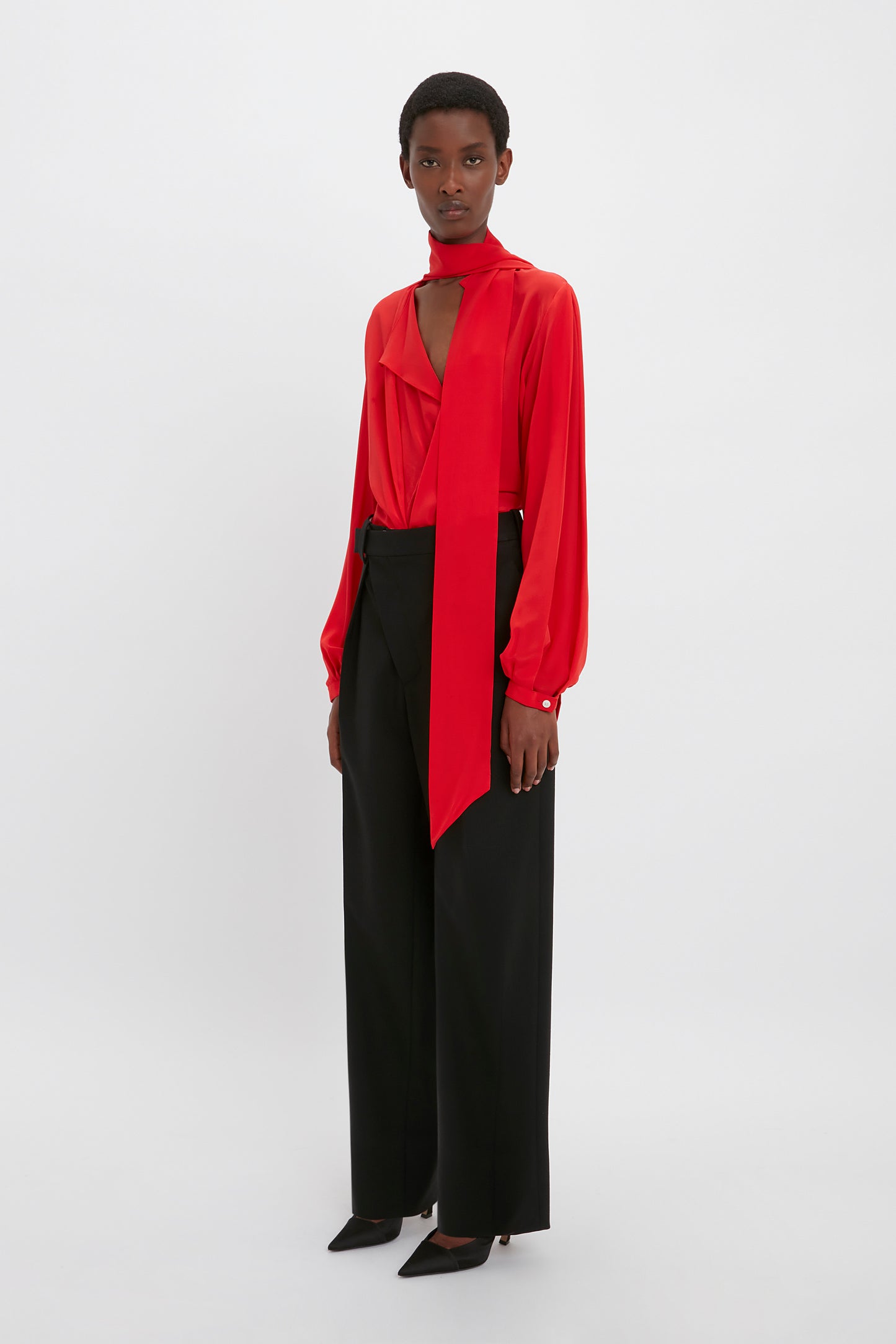 A person stands against a plain background, wearing a bright red Victoria Beckham Scarf Neck Blouse In Bright Red with a sophisticated deep-V neckline and long tied scarf. The ensemble is completed with black high-waisted pants and black pointed-toe shoes.