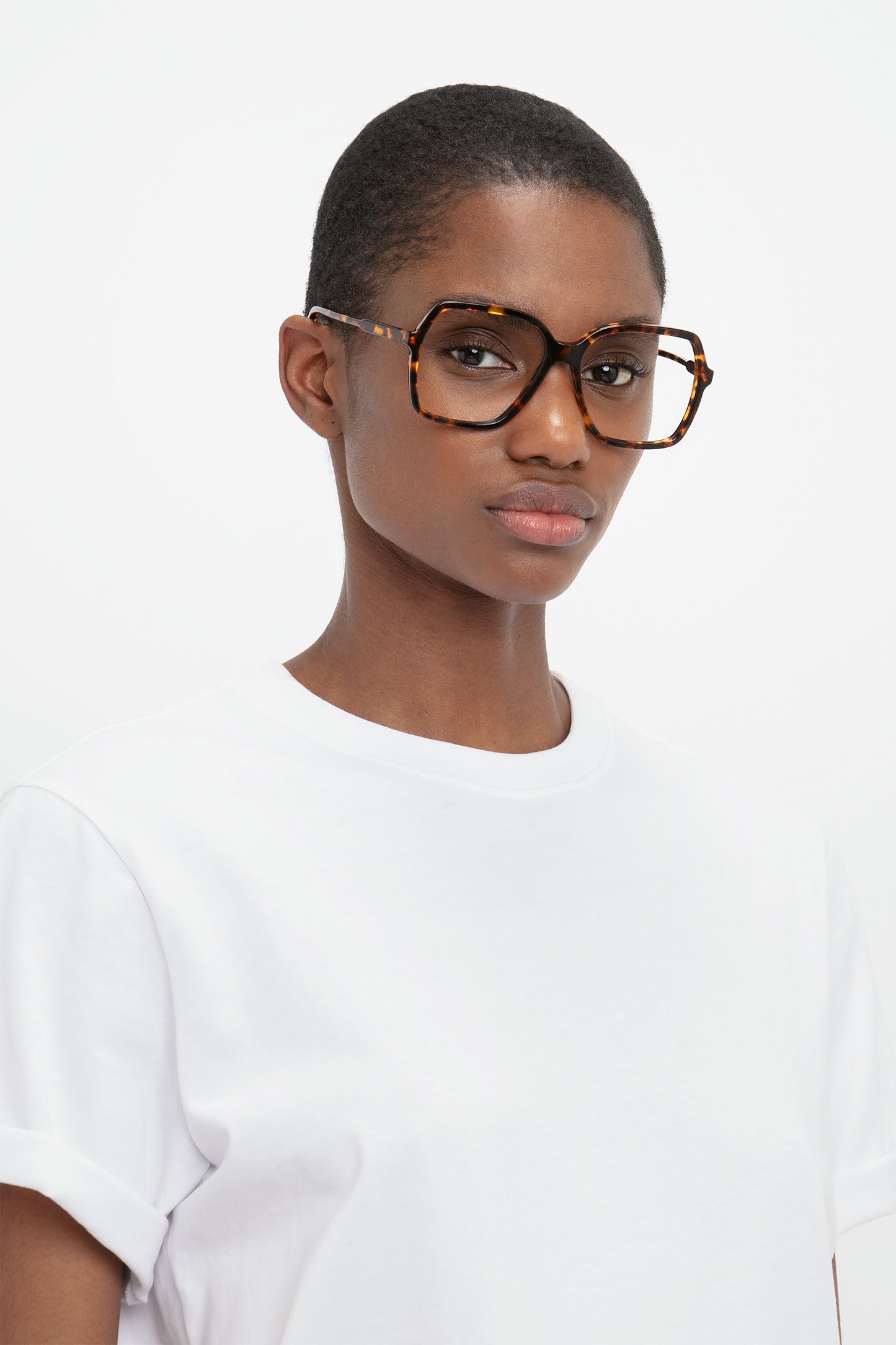 A person with short hair wearing Guilloche Frame Opticals In Dark Havana from the Victoria Beckham collection and a white t-shirt is looking at the camera against a plain background.