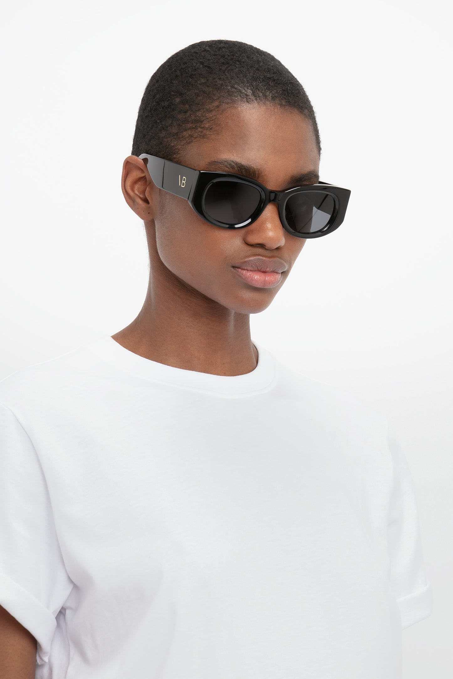 A person wearing black rectangular Monogram Detail Sunglasses In Black by Victoria Beckham and a white T-shirt stands against a plain white background.