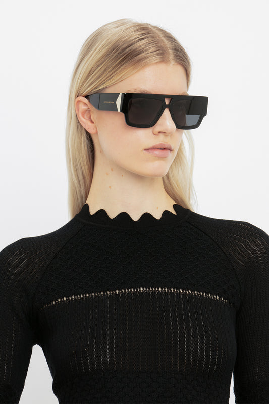 Woman with blonde hair wearing futuristic black Victoria Beckham V Plaque Frame Sunglasses with a double bridge, and a black scalloped-neckline top, against a white background.