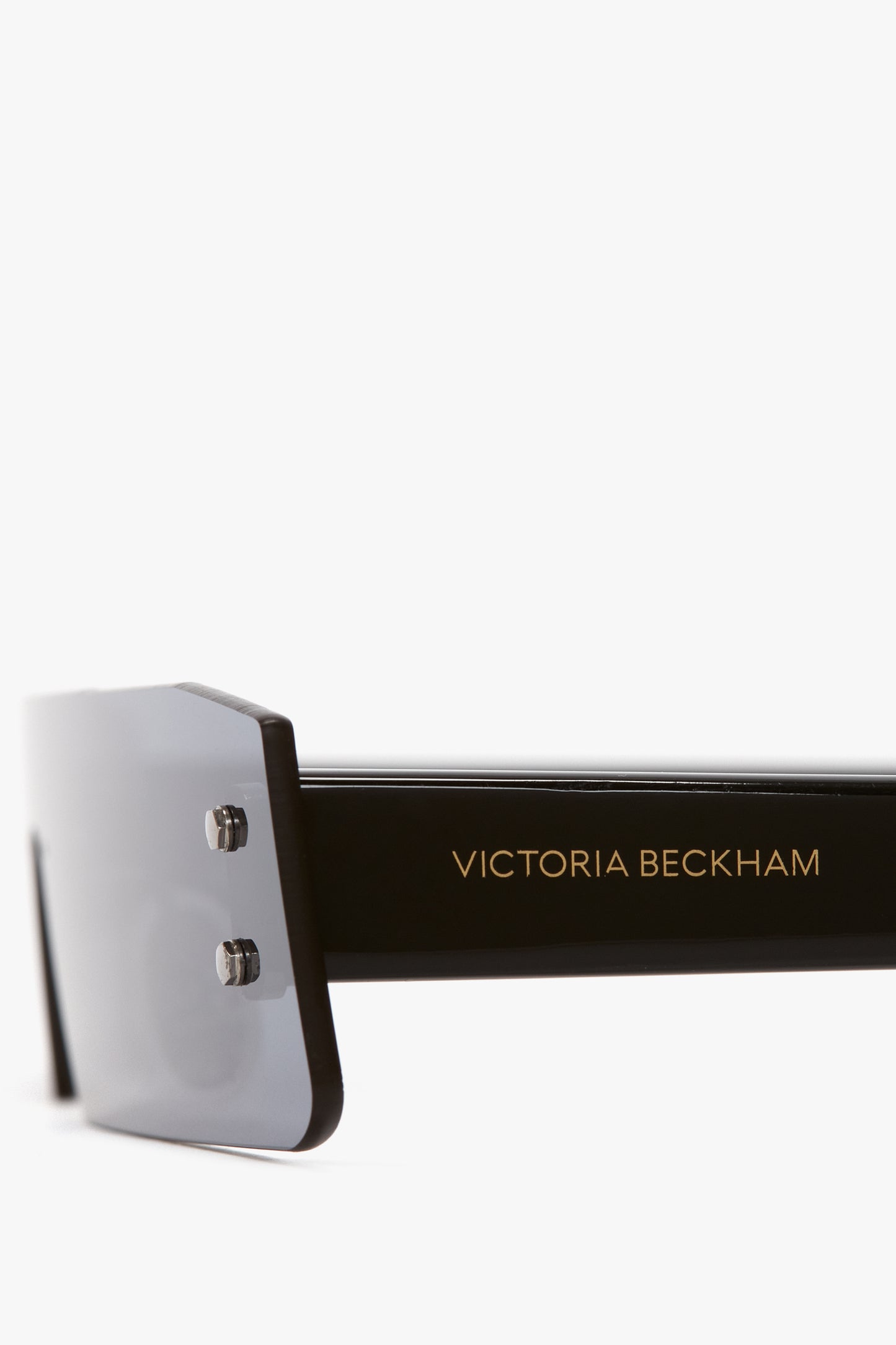 Close-up of black sunglasses with a thick frame and dark blue mirror lenses. The arm of the sunglasses displays the gold text "Victoria Beckham.