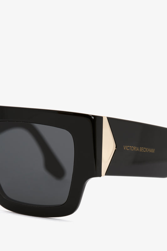 Close-up of a black rectangular Victoria Beckham V Plaque Frame sunglasses with gold accents, isolated on a white background.