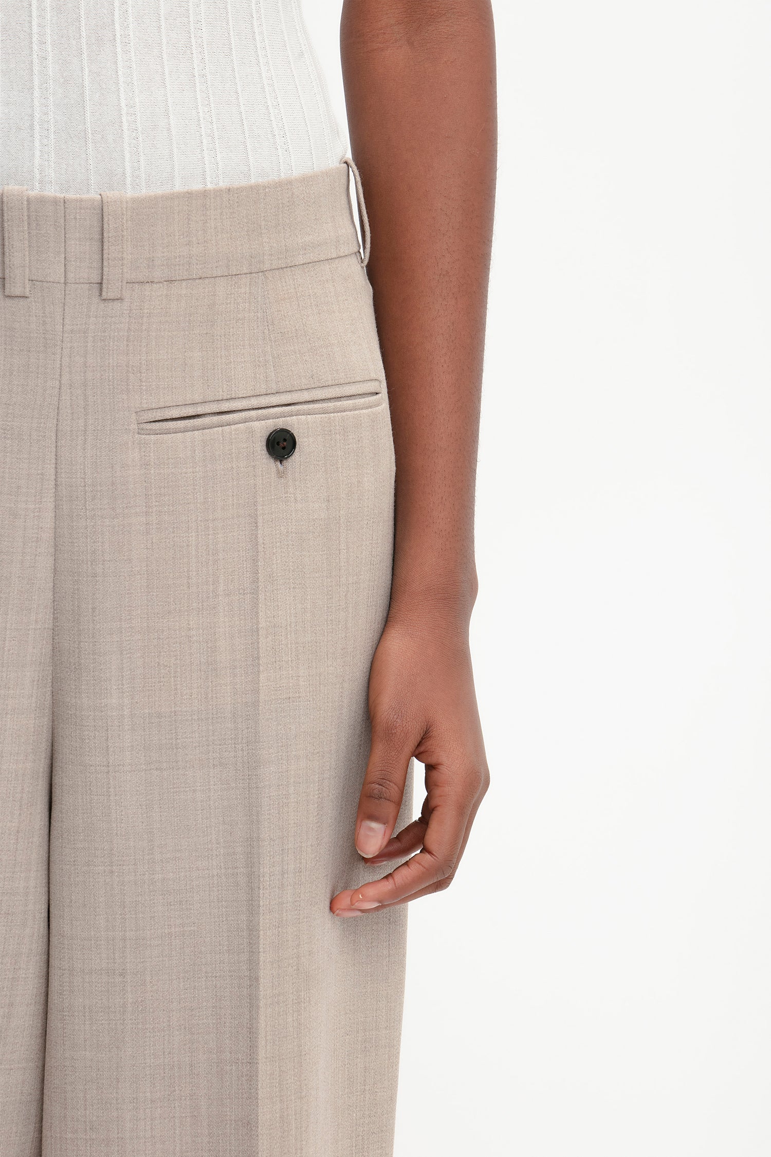 Close-up of a modern woman's arm resting alongside her body, wearing Victoria Beckham Reverse Front Trouser In Sesame with a single black button on a small pocket, and a white shirt tucked into the waistband.