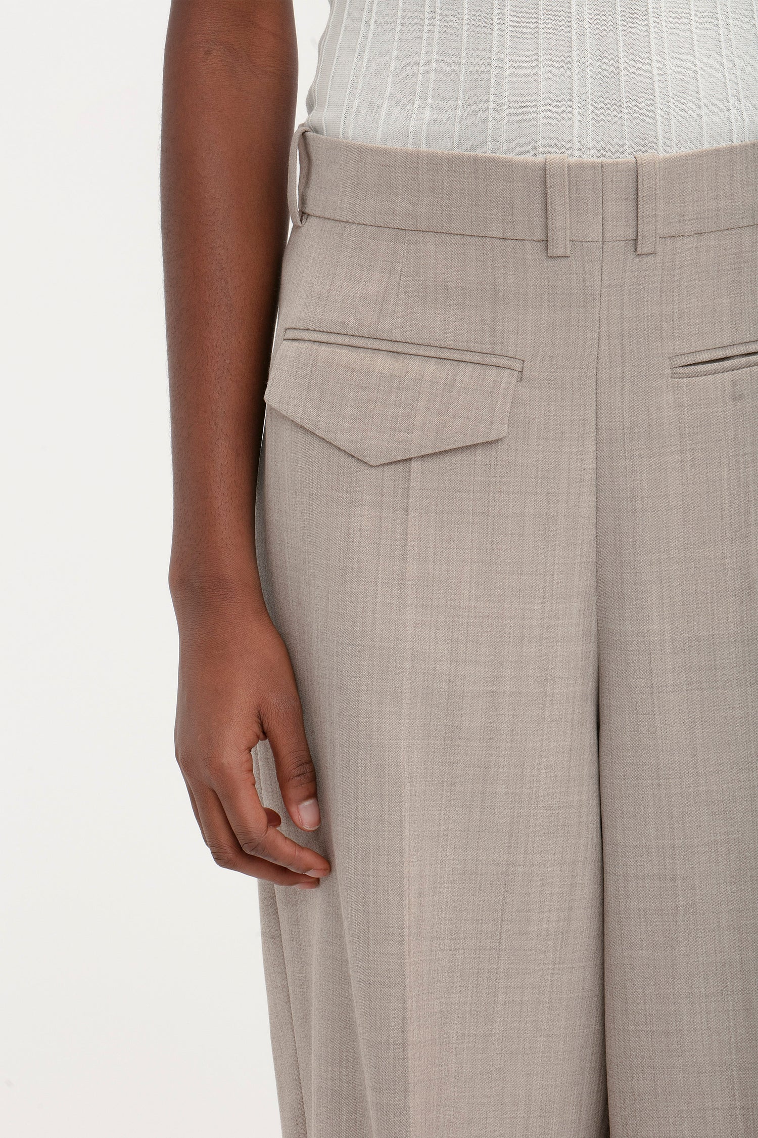 A modern woman wearing a white ribbed top and beige Victoria Beckham Reverse Front Trouser In Sesame with flap pockets, stands with her right arm relaxed at her side, showcasing versatile styling.