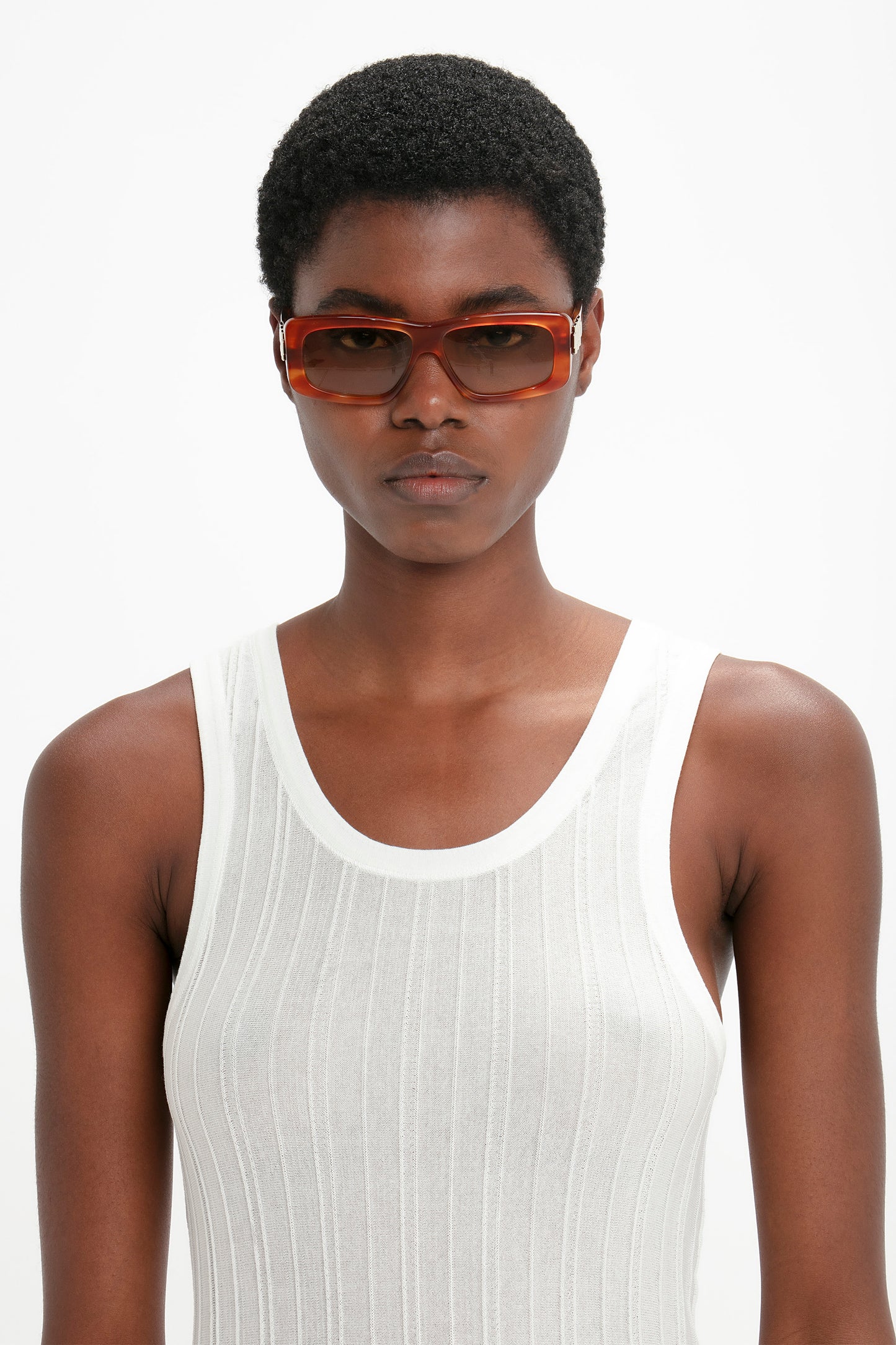 Person with short hair wearing a white sleeveless top and Chain Detail Rectangular Frame Sunglasses In Striped Blonde Havana from Victoria Beckham, standing against a plain white background.