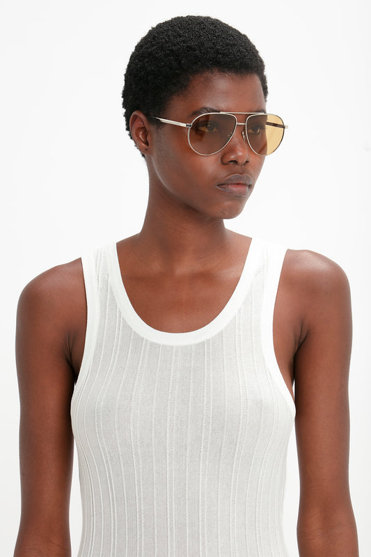 A young woman with short hair wearing a white tank top and Victoria Beckham's V Metal Pilot Sunglasses In Silver-Brown with adjustable nose pads, facing slightly to her right in front of a white background.