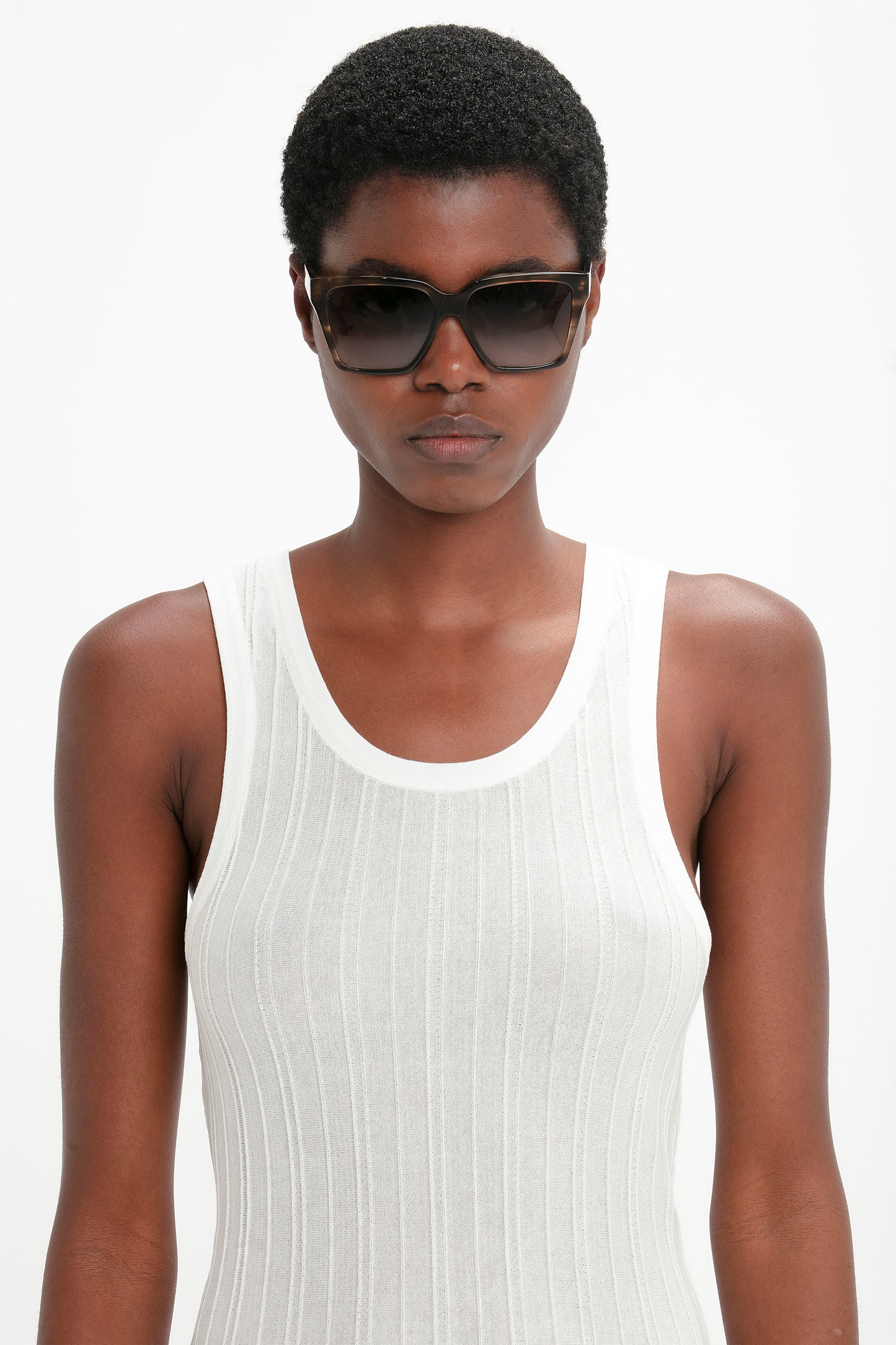 A person wearing dark sunglasses and a white sleeveless ribbed shirt against a plain background, showcasing stylish Victoria Beckham Soft Square Frame Sunglasses In Striped Khaki Havana.
