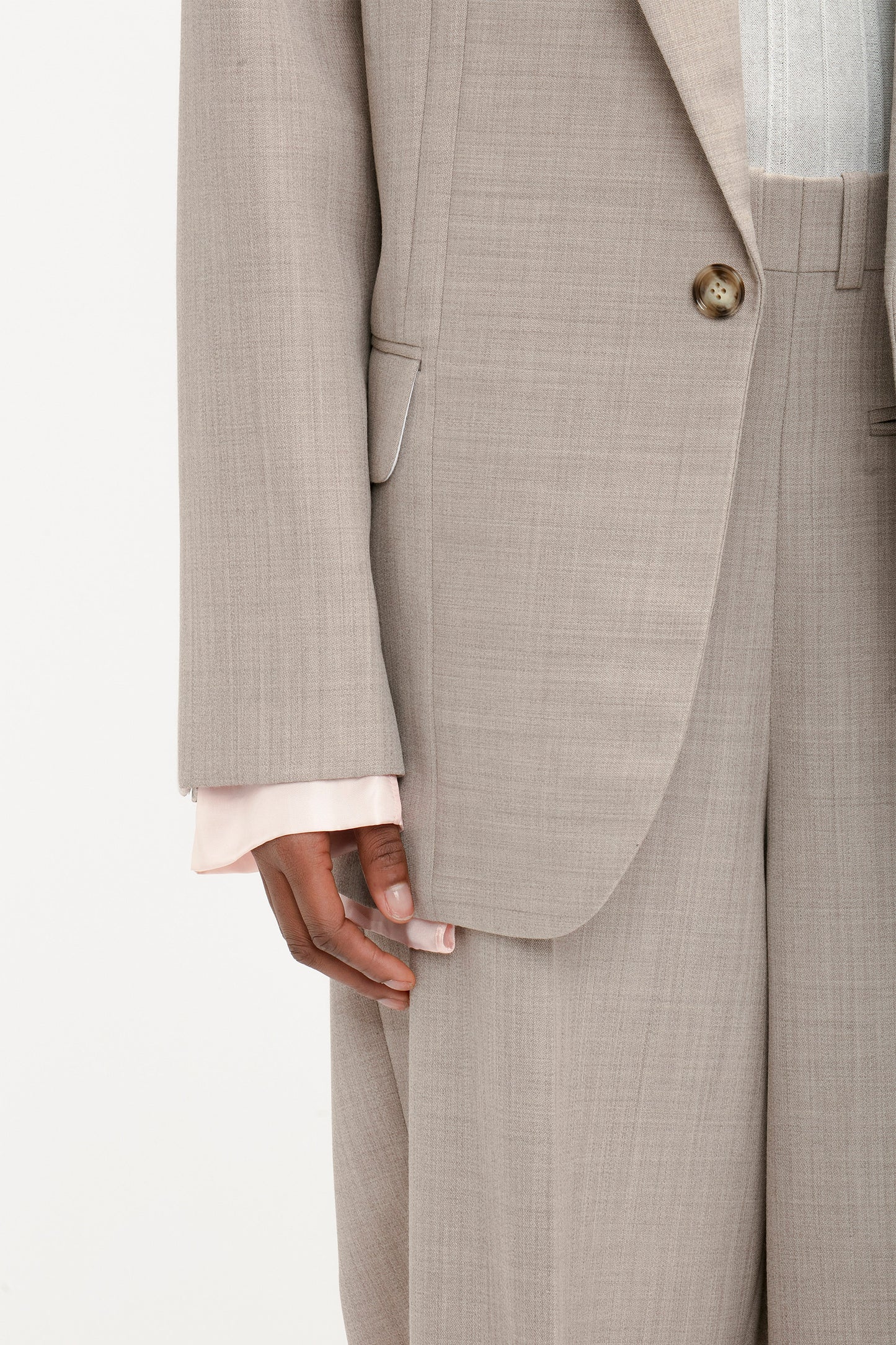 A person wearing a Victoria Beckham Darted Sleeve Tailored Jacket In Sesame, with contemporary detailing visible, paired with beige pants and a light pink shirt sleeve peeking out from under the jacket sleeve.