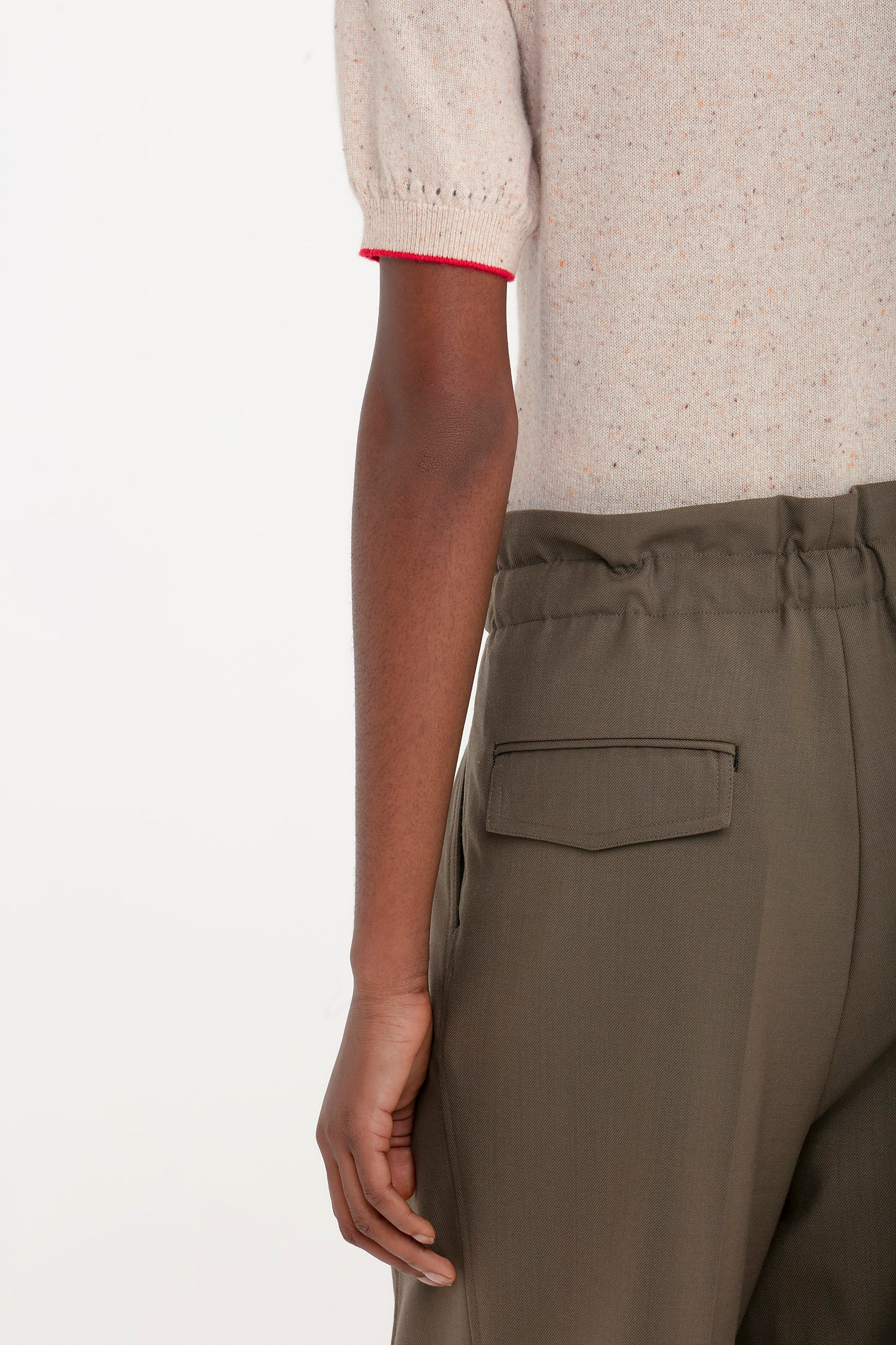 A person wearing olive green pants with a back pocket and an off-white Short Sleeve Top In Nougat by Victoria Beckham with red stitching is shown from behind. Their left hand is visible beside their body, accentuating the lightweight cashmere knit of the top with its feminine punctuation.