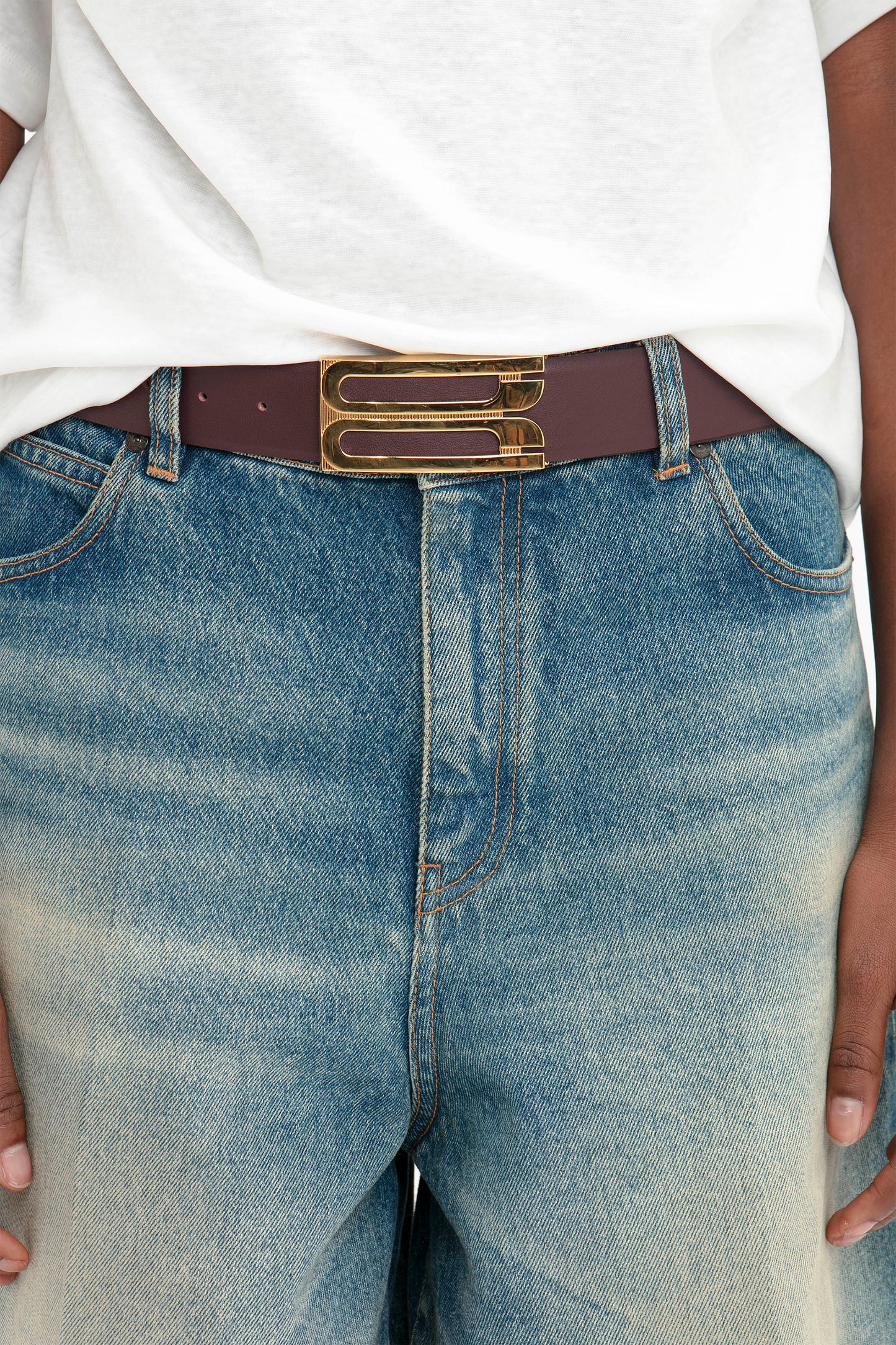 A person wears the Victoria Beckham Oversized Bermuda Short In Antique Indigo Wash with a white shirt, accessorized with a brown belt featuring a distinctive double buckle.