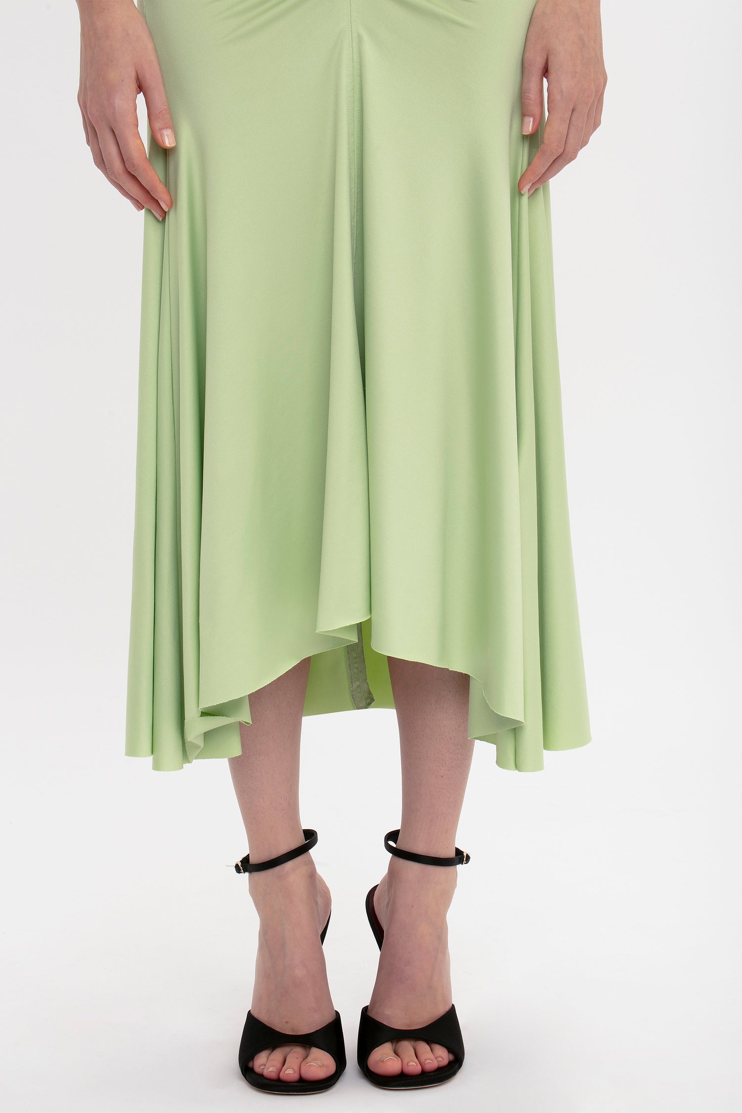 Woman standing in a Victoria Beckham sleeveless ruched jersey dress in pistachio and black ankle-strap heels, with her hands gently resting on her hips.