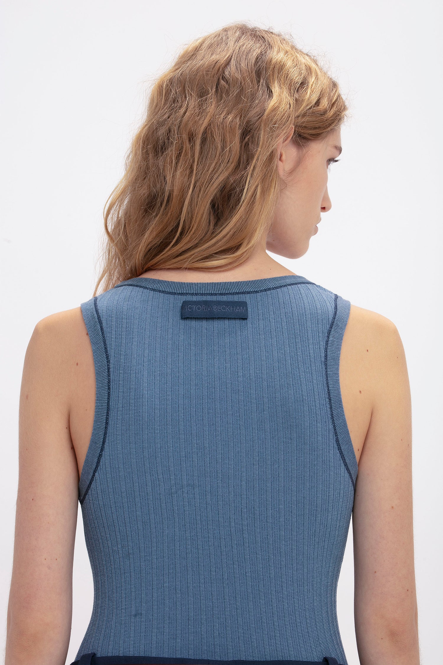 A person with long, wavy hair is facing away, wearing a sleeveless, ribbed *Fine Knit Micro Stripe Tank In Heritage Blue* by *Victoria Beckham* against a plain background, reminiscent of Victoria Beckham's classic style.