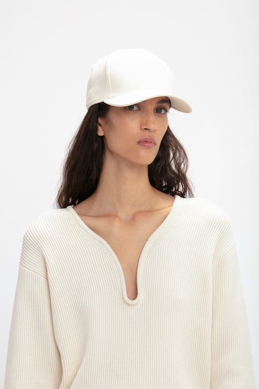 A woman in a Victoria Beckham logo cap in antique white and cream sweater looks directly at the camera with a neutral expression.