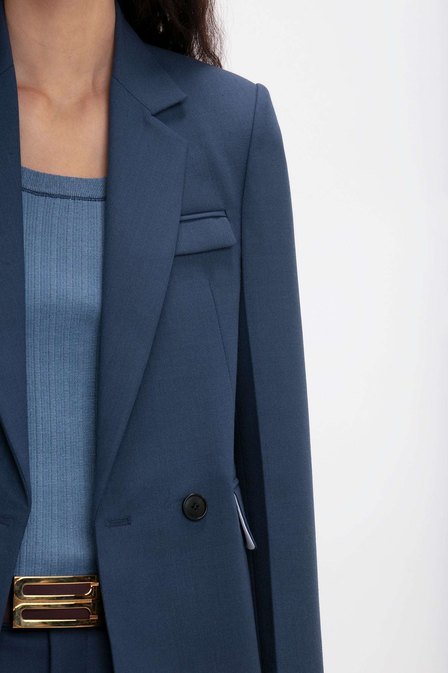 Close-up of a woman wearing a Victoria Beckham Shrunken Double Breasted Jacket in Heritage Blue over a ribbed top, with a partial view of a gold belt.