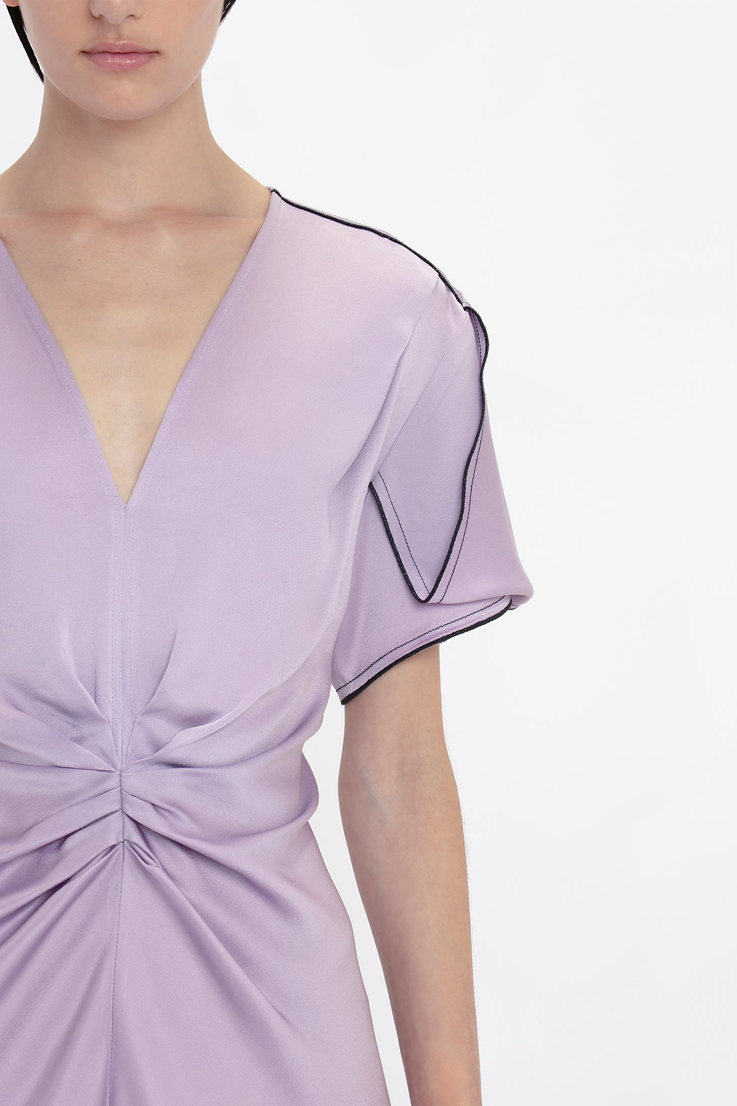 A person in an elegant Gathered V-Neck Midi Dress In Petunia with a deep V-neckline, gathered detail at the waist, and black trim along the edge of the sleeve, photographed against a plain white background. This piece exudes sophistication reminiscent of Victoria Beckham's designs.