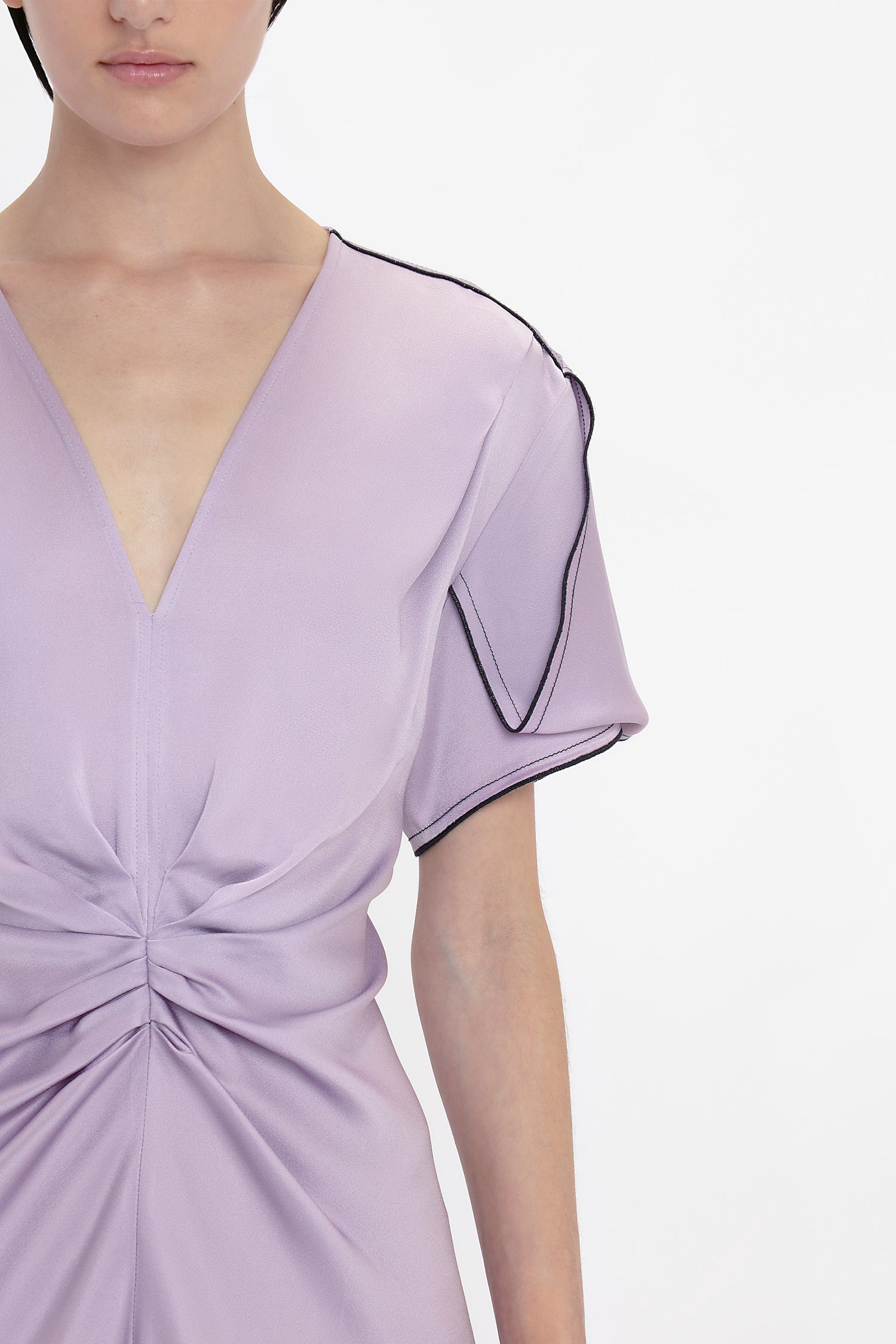 A person in an elegant Gathered V-Neck Midi Dress In Petunia with a deep V-neckline, gathered detail at the waist, and black trim along the edge of the sleeve, photographed against a plain white background. This piece exudes sophistication reminiscent of Victoria Beckham's designs.