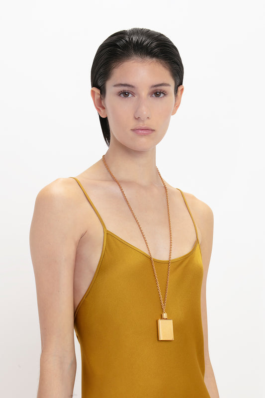 Young woman in a Victoria Beckham Low Back Cami Floor-Length Dress In Harvest Gold with a long necklace, standing against a white background, looking directly at the camera.