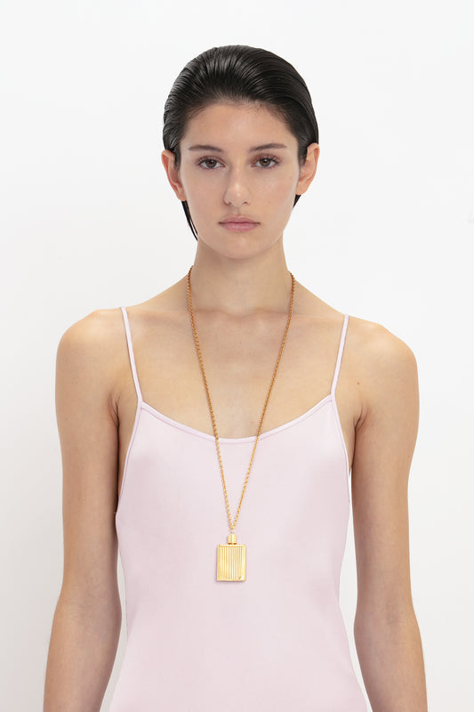 A young woman with slicked-back hair wearing a Victoria Beckham light pink camisole and a large gold pendant necklace, standing against a white background.