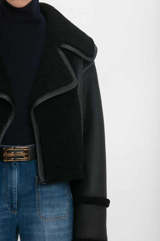 Close-up of a person wearing a dark turtleneck, Victoria Beckham shearling jacket in black, and jeans, with a focus on the jacket and belt details.
