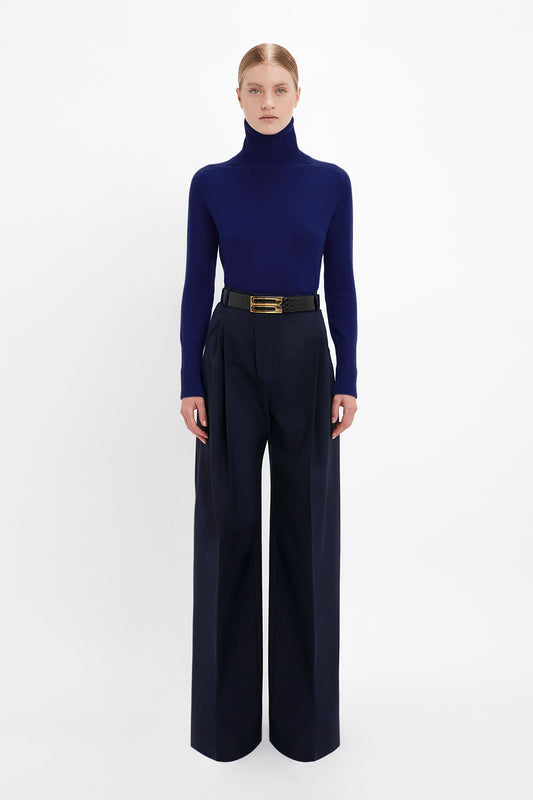 A person stands against a plain white background, wearing a dark blue turtleneck sweater and Wide Leg Trouser In Midnight by Victoria Beckham, secured with a black belt.