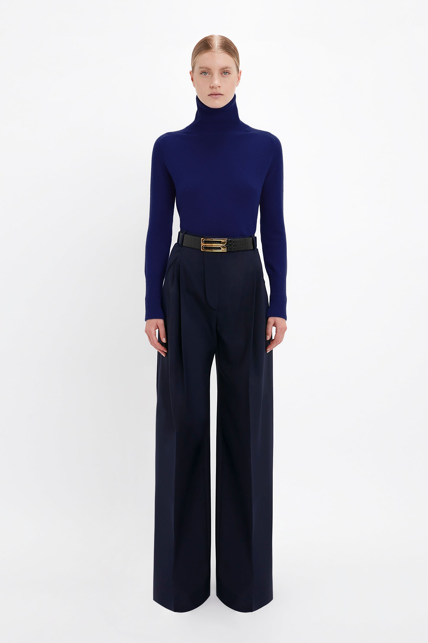A person stands against a plain white background wearing a dark blue textured knit Polo Neck Jumper In Navy by Victoria Beckham, black belt, and wide-legged navy trousers.