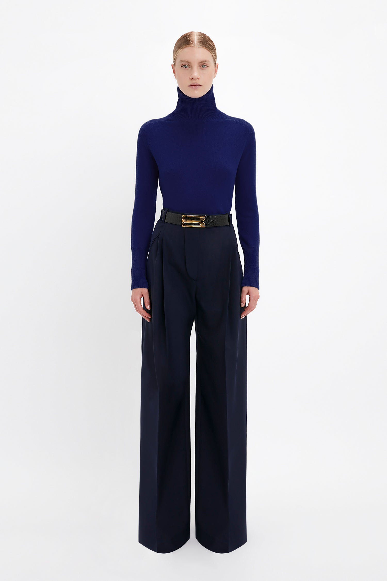 A person stands against a white background wearing a Victoria Beckham Polo Neck Jumper In Navy and wide-leg dark pants, accessorized with a belt.