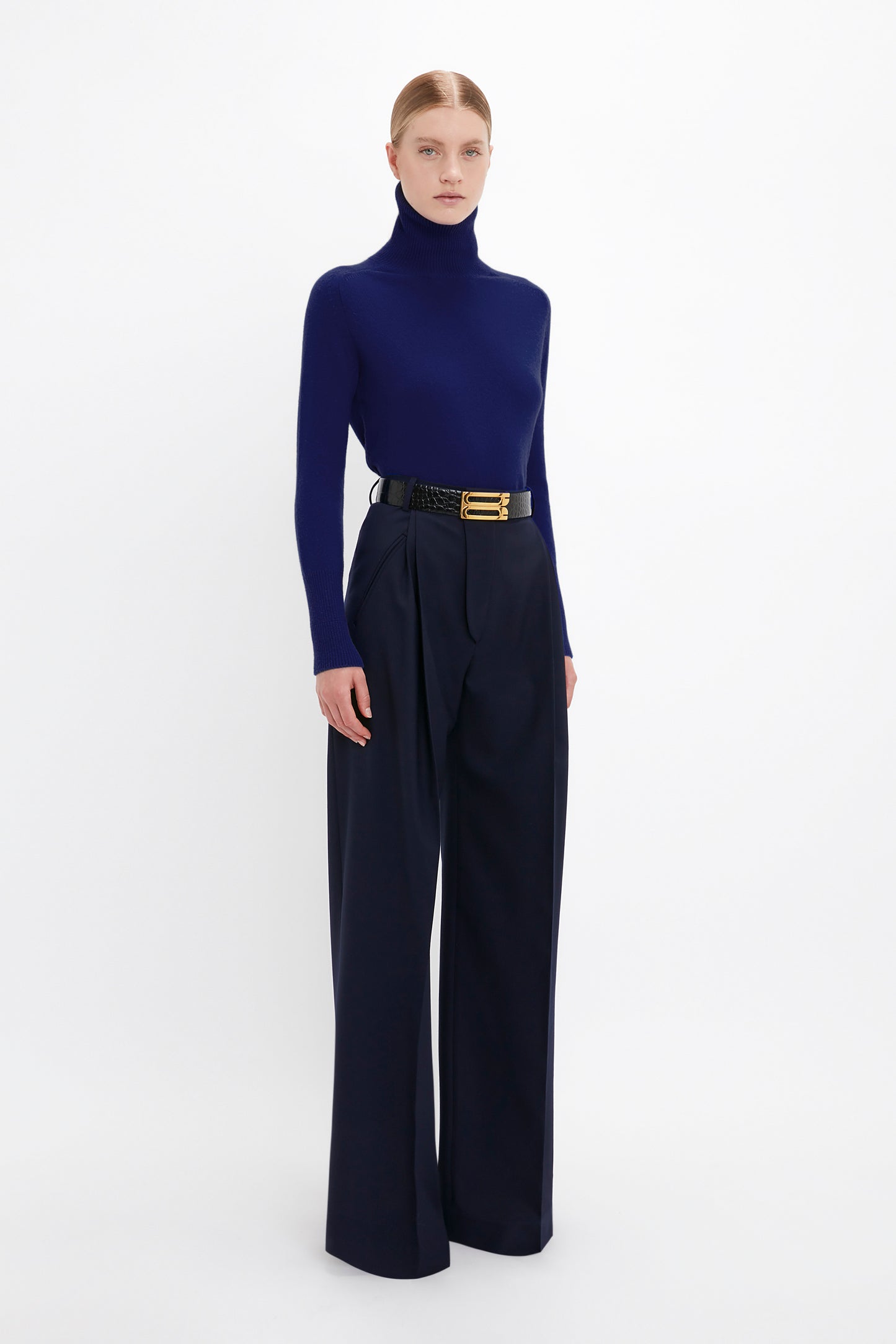 A person is standing and wearing a Polo Neck Jumper In Navy by Victoria Beckham with high-waisted wide-leg trousers, and a black belt with a gold buckle. The background is plain white.