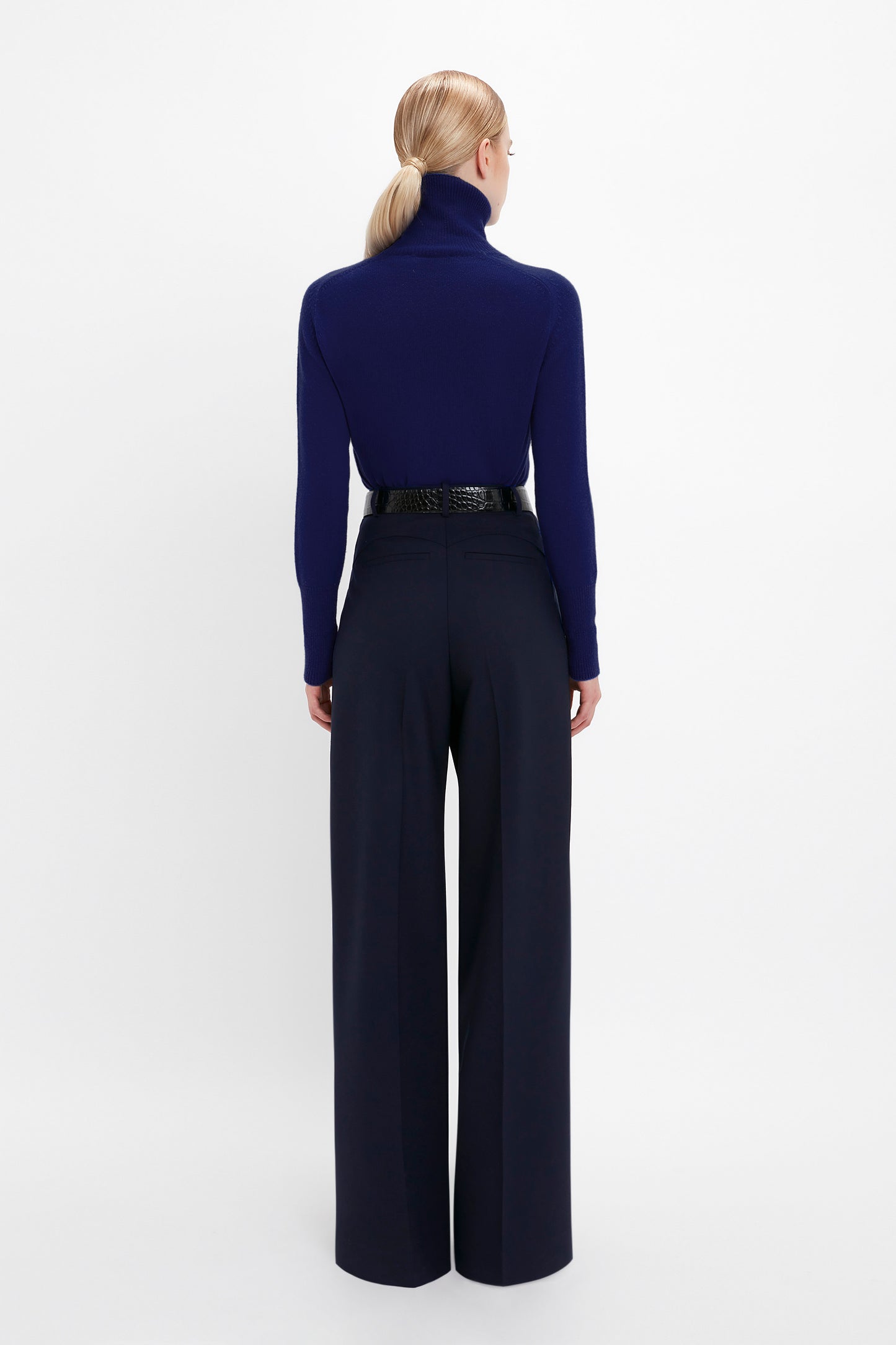 Person with blonde hair in a ponytail, wearing a Polo Neck Jumper In Navy by Victoria Beckham featuring a contrasting embroidered monogram, dark wide-leg pants, and a black belt, facing away from the camera against a white background.