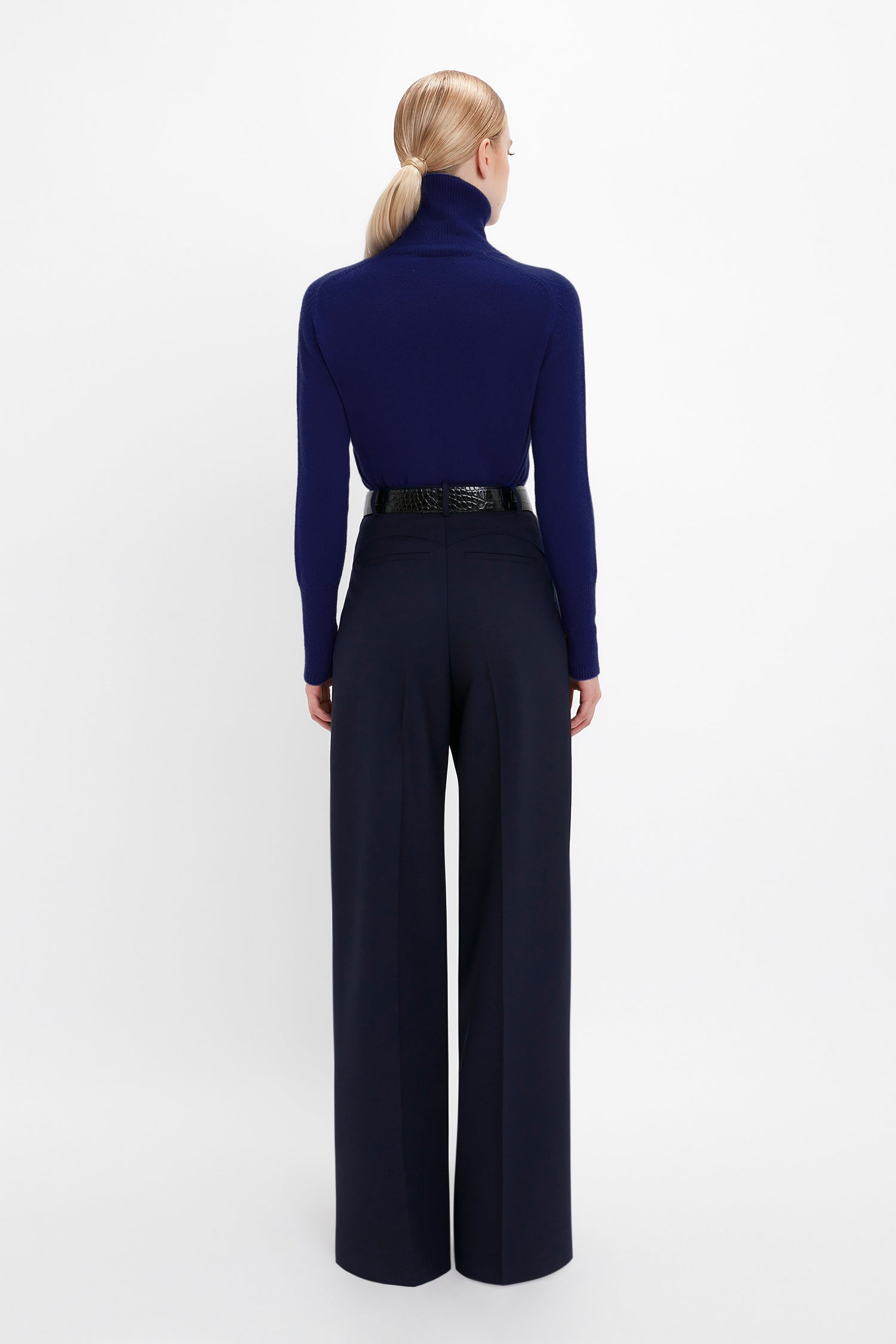 Person with blonde hair in a ponytail, wearing a Polo Neck Jumper In Navy by Victoria Beckham featuring a contrasting embroidered monogram, dark wide-leg pants, and a black belt, facing away from the camera against a white background.