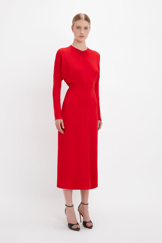 A woman in a plain red Victoria Beckham Dolman Midi Dress stands against a white background, paired with black pointy toe stiletto heels.