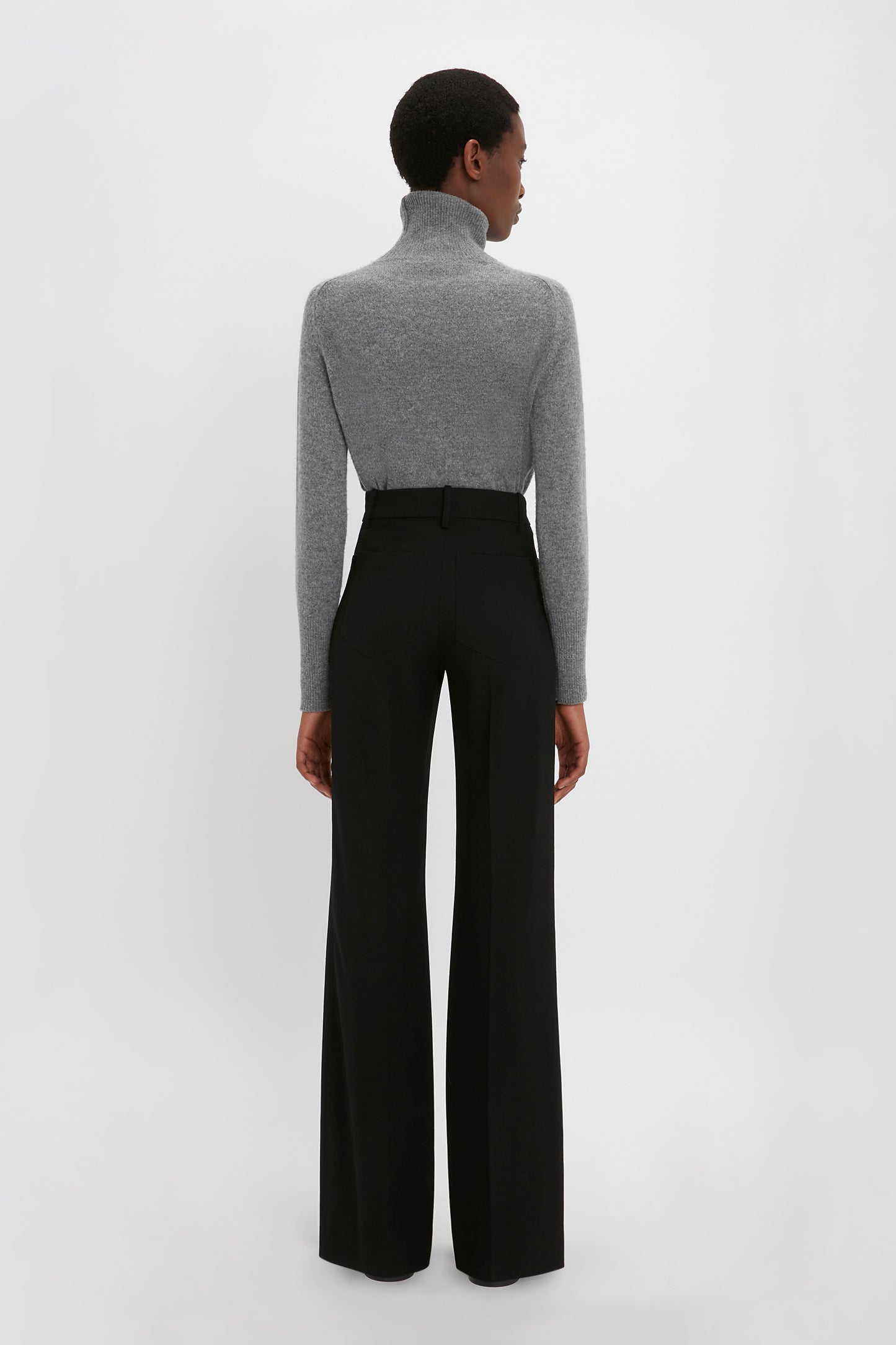Rear view of a woman standing against a white background, wearing a gray turtleneck and Victoria Beckham's Alina Tailored Trouser In Black.