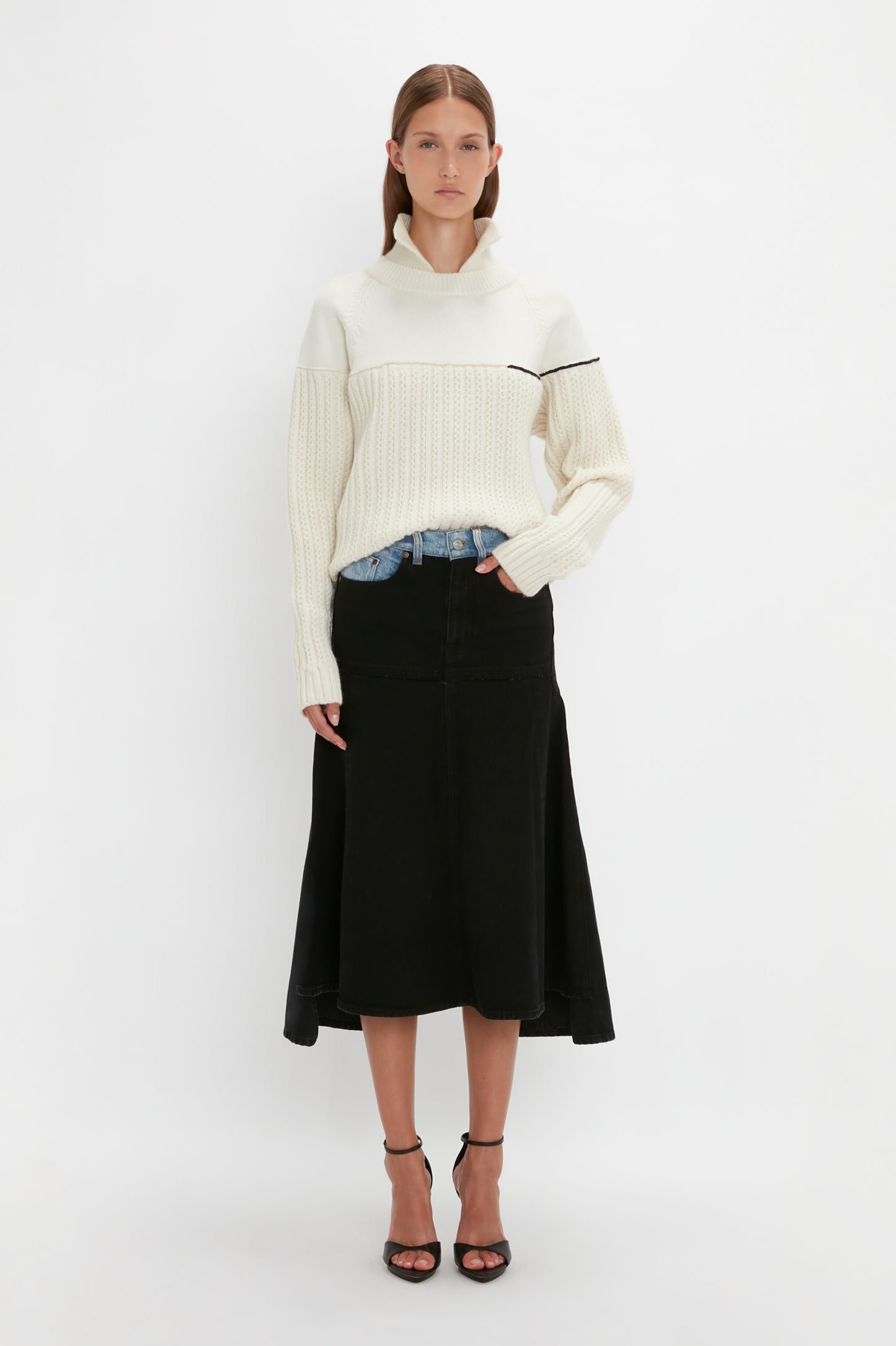 A person stands against a plain background wearing the Collar Detail Jumper In Natural by Victoria Beckham with casual sophistication, complemented by a black skirt and black heels.