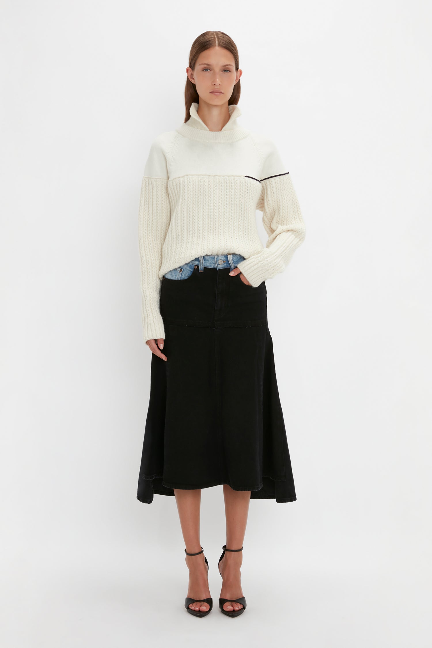 A person stands against a plain background wearing the Collar Detail Jumper In Natural by Victoria Beckham with casual sophistication, complemented by a black skirt and black heels.