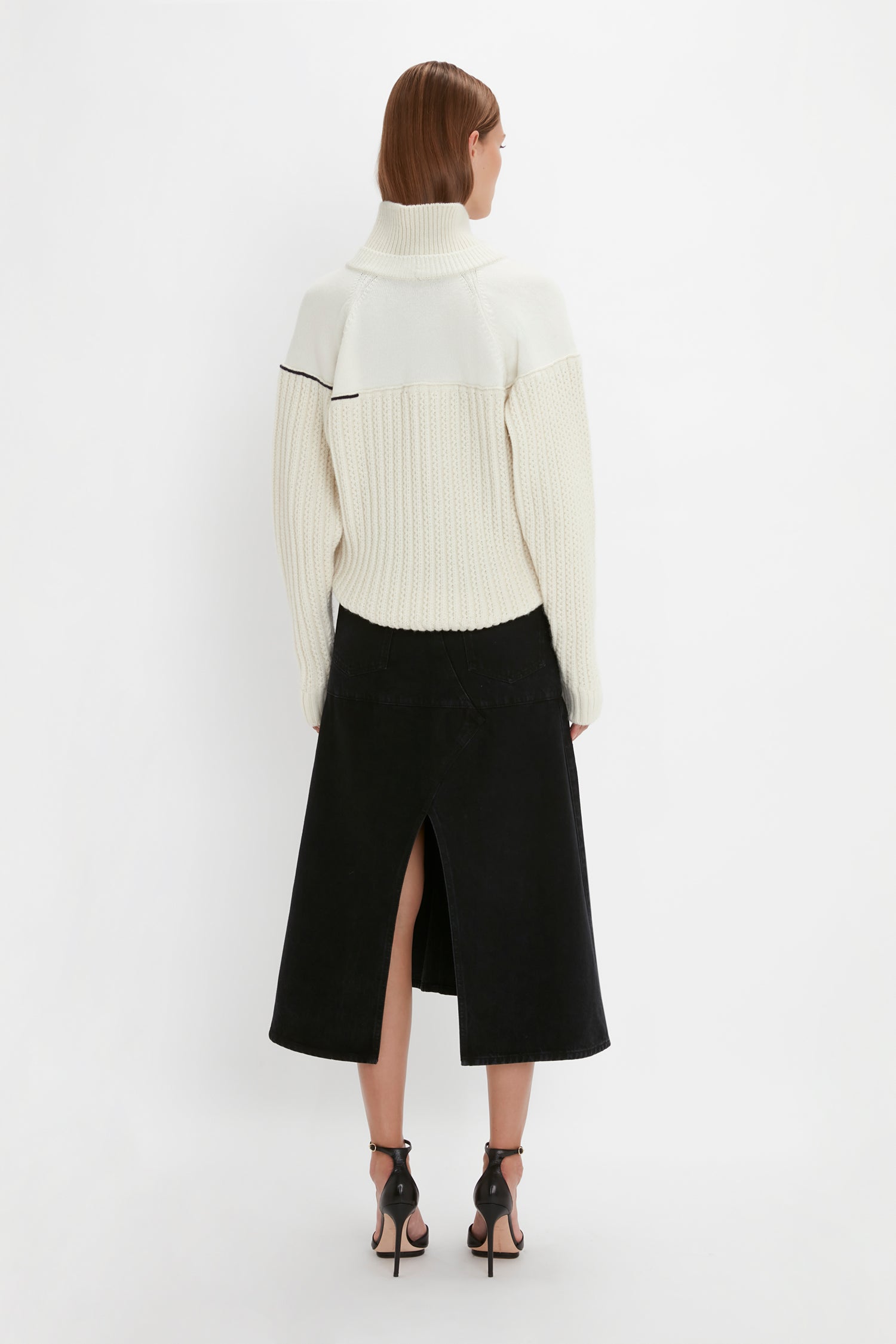 A person wearing a white Victoria Beckham Collar Detail Jumper In Natural, a black skirt with a back slit, and high-heeled sandals stands against a plain white background, facing away from the camera.