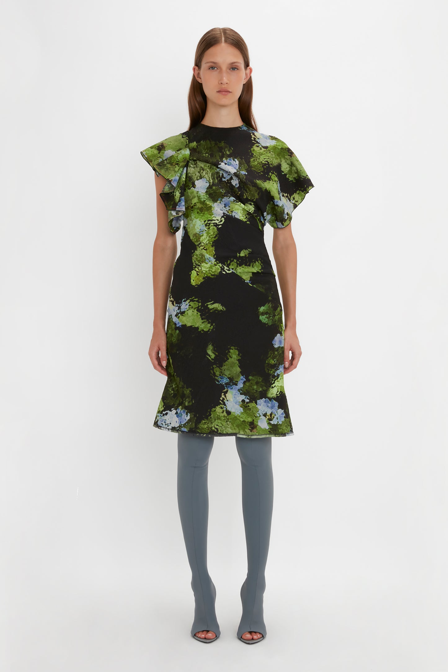 A person stands against a white background wearing the Twist Shoulder Dress In Black Frost by Victoria Beckham, complemented by gray knee-high boots.