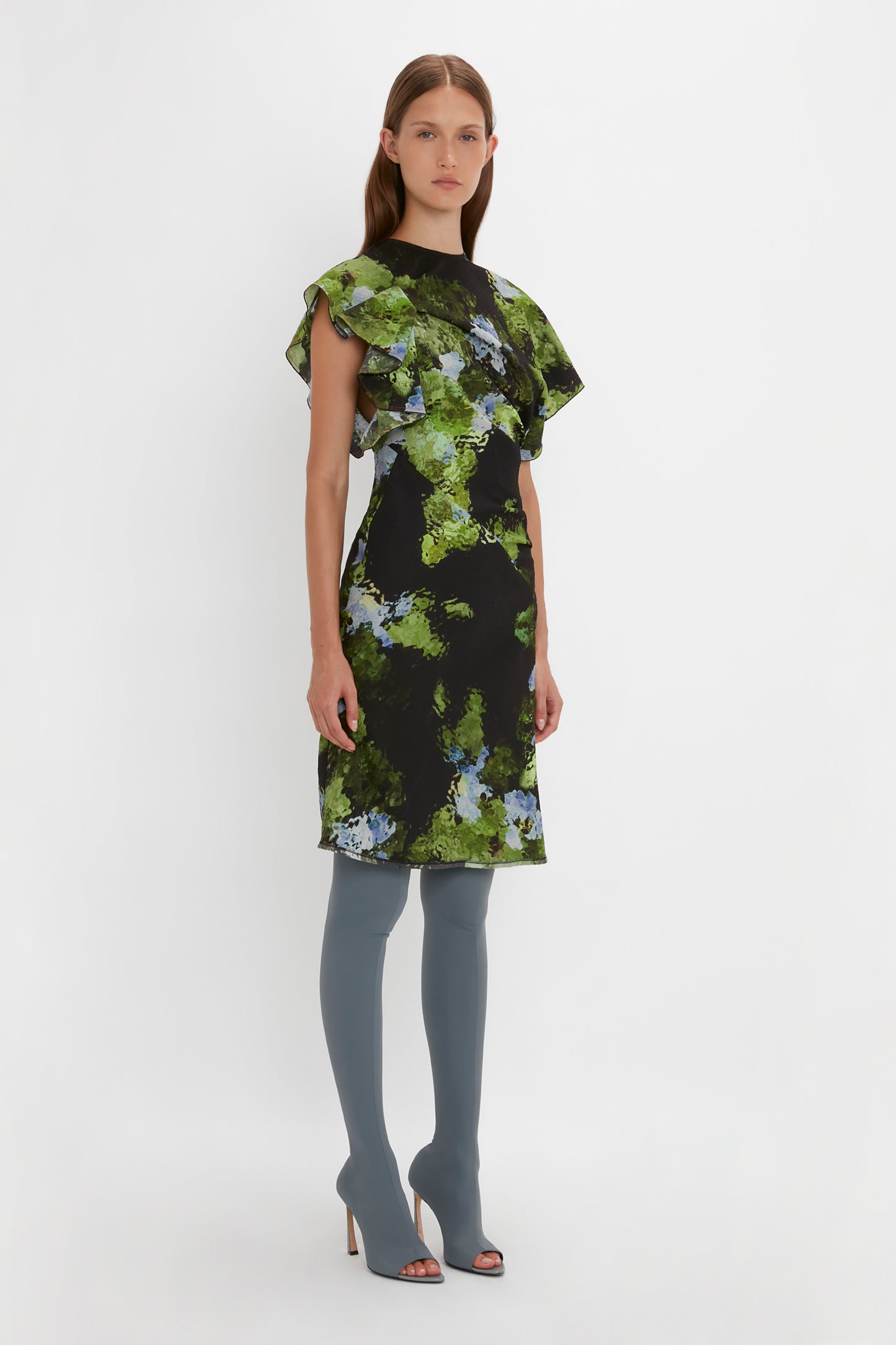 A person stands facing forward wearing a black Twist Shoulder Dress In Black Frost by Victoria Beckham with green and blue floral patterns, featuring an asymmetric hemline and waist-defining fit, paired with gray thigh-high boots against a white background.