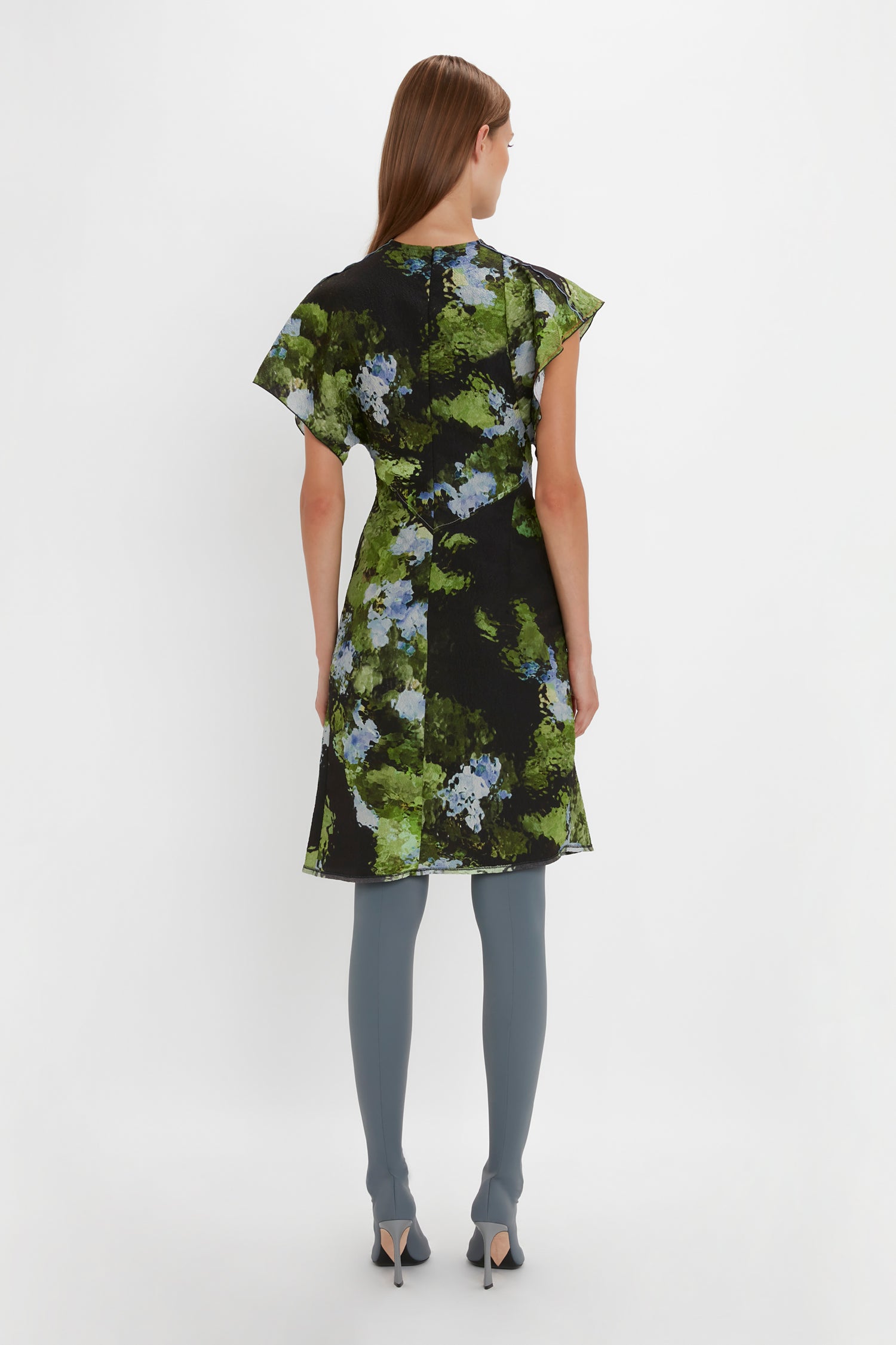A person is standing with their back to the camera, showcasing a Victoria Beckham Twist Shoulder Dress In Black Frost adorned with green and blue floral patterns. Complementing the waist-defining fit, they pair it with blue stockings and black high-heeled shoes.